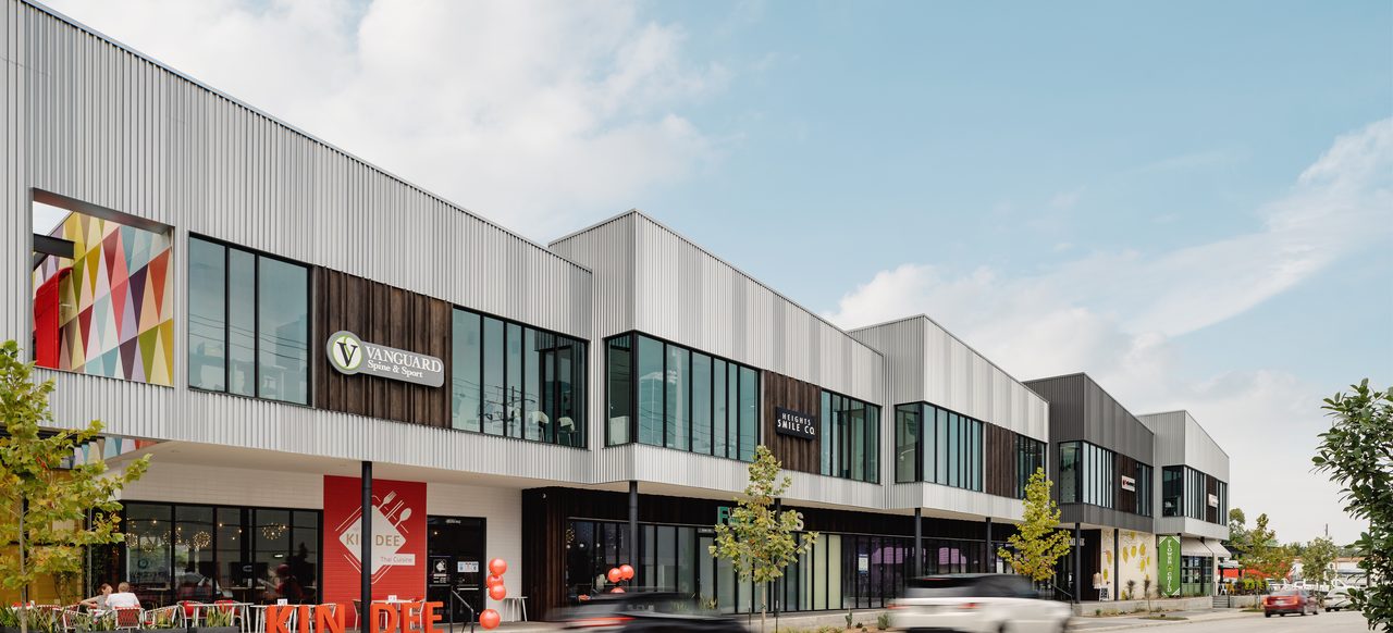 The two-story storefront row at Market at Houston Heights along North Shepherd Drive — Kin Dee, Vanguard Spine & Sport and Heights Smile Co. above the ground-floor restaurants, with traffic passing in the foreground